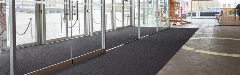 Wide entrance mat at the front of a UK office building with people walking in — designed to reduce dirt and slips, featuring a clean modern look